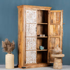 Side angle image with open door of a mango wood wardrobe with whitewashed floral carvings, vintage furniture cabinet in carved wood, 190 × 105 × 42 cm.