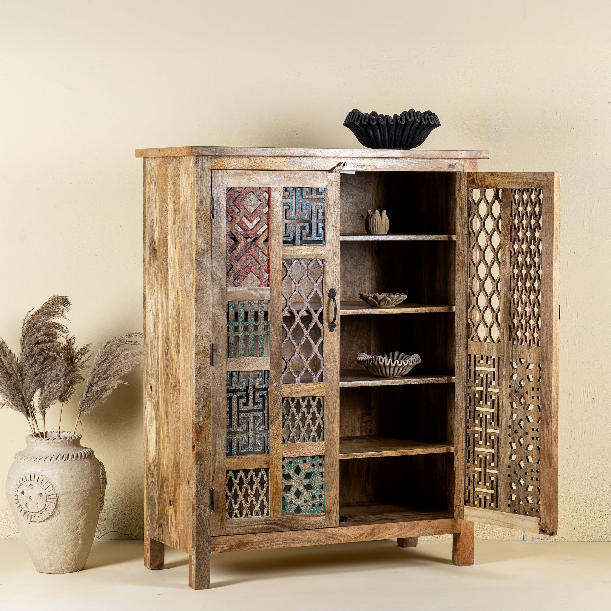 Side angle with open door image for a   artisan-made cabinet crafted from mango wood, featuring double doors adorned with a patchwork of colourful, intricately carved geometric panels, blending traditional craftsmanship with a vibrant, eclectic style.
