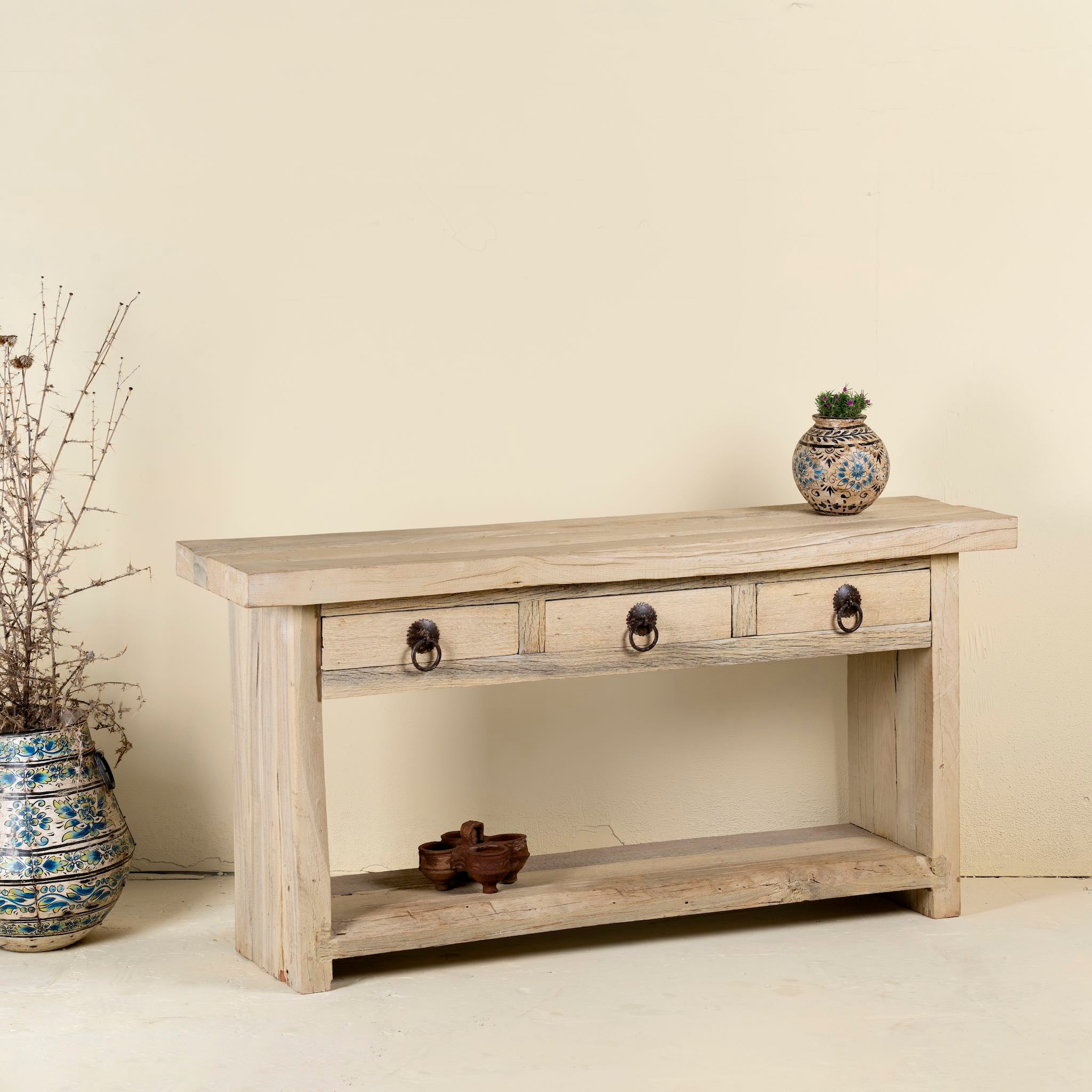 White background image of a rustic console table crafted from reclaimed teak wood, featuring a natural weathered finish, three drawers with metal ring pulls, and a lower shelf for additional storage or display, blending simplicity with organic charm.

