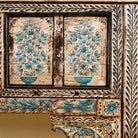 Close up image of a  hand-painted console table made from reclaimed teak wood, featuring intricate floral motifs in blue tones across the front panels, framed by carved detailing and a distressed finish that highlights its artisanal and vintage character.
