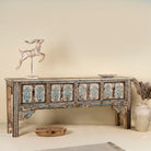Side angle image of a hand-painted console table made from reclaimed teak wood, featuring intricate floral motifs in blue tones across the front panels, framed by carved detailing and a distressed finish that highlights its artisanal and vintage character.
