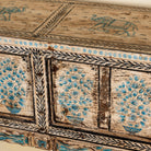 Close up side image of a hand-painted console table made from reclaimed teak wood, featuring intricate floral motifs in blue tones across the front panels, framed by carved detailing and a distressed finish that highlights its artisanal and vintage character.
