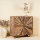 Side angle image of a artistic wooden cabinet in rustic style, made from reclaimed wood with a striking sunburst pattern on the double doors, featuring subtle color variations and round metal ring pulls, elevated on rounded wooden feet.

