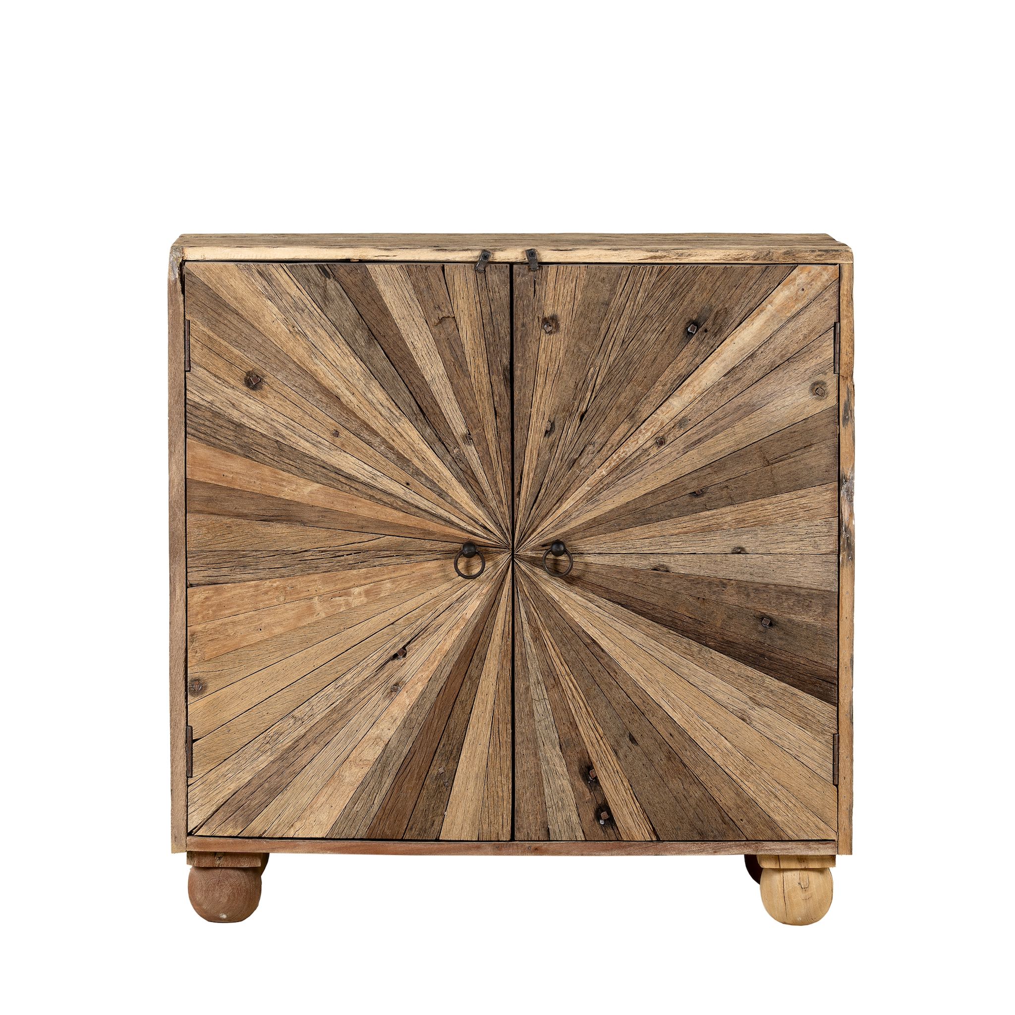 White background image of a artistic wooden cabinet in rustic style, made from reclaimed wood with a striking sunburst pattern on the double doors, featuring subtle color variations and round metal ring pulls, elevated on rounded wooden feet.
