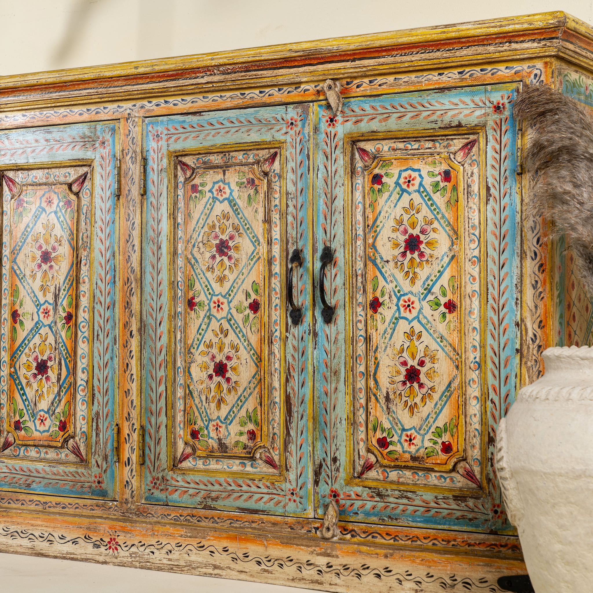 Side angle close up image of a hand-painted sideboard made from reclaimed wood, featuring four intricately detailed doors with vibrant floral and geometric motifs in soft blues, yellows, and reds, finished with a distressed vintage patina for a charming, artisanal look.
