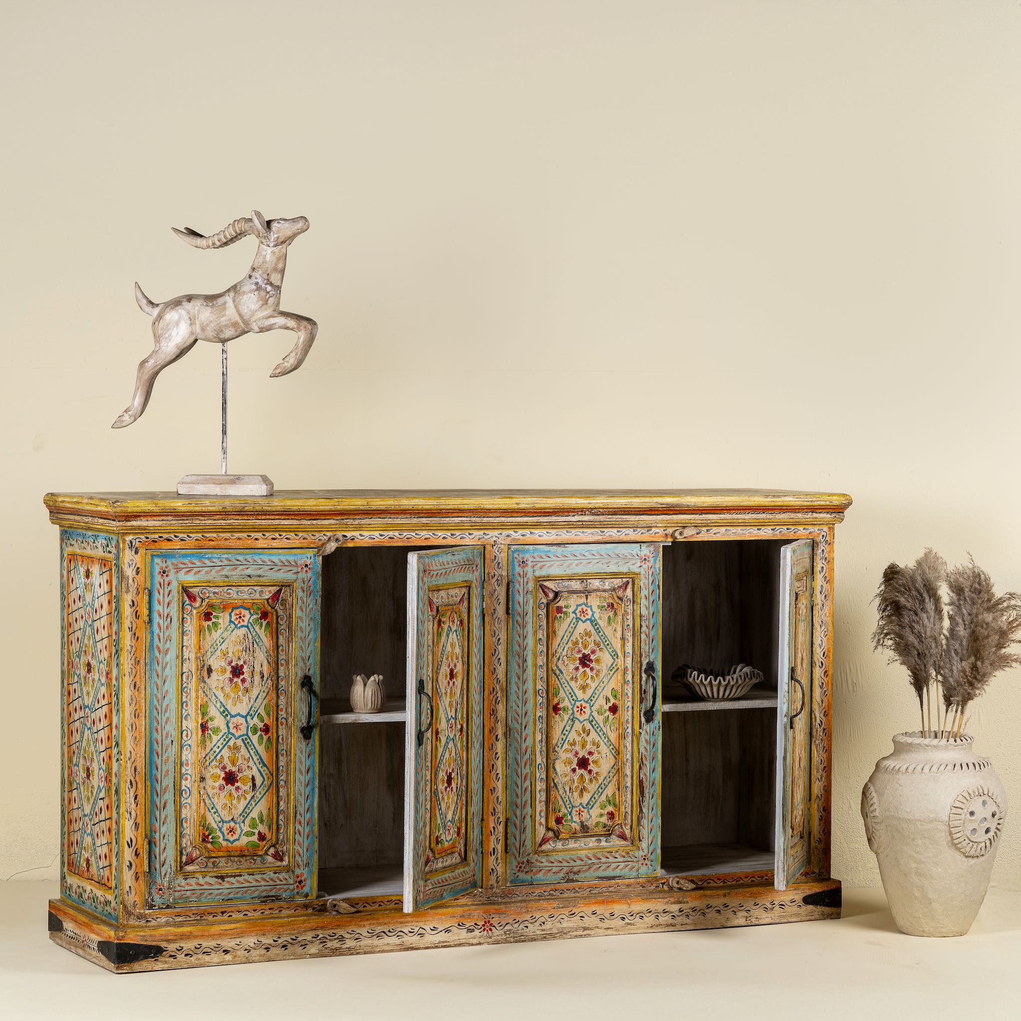 Side angle image with open doors of a hand-painted sideboard made from reclaimed wood, featuring four intricately detailed doors with vibrant floral and geometric motifs in soft blues, yellows, and reds, finished with a distressed vintage patina for a charming, artisanal look.
