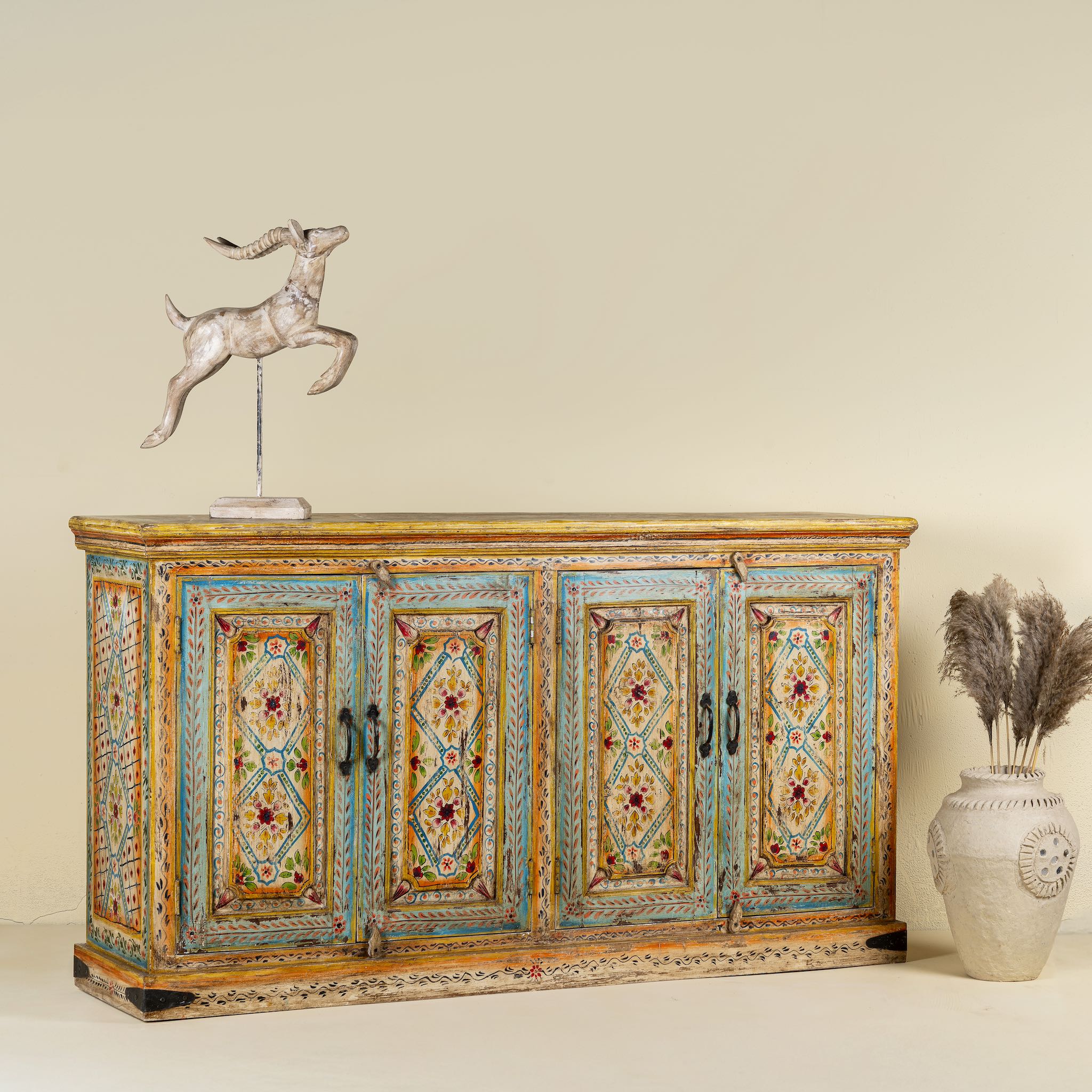 Side angle image of a hand-painted sideboard made from reclaimed wood, featuring four intricately detailed doors with vibrant floral and geometric motifs in soft blues, yellows, and reds, finished with a distressed vintage patina for a charming, artisanal look.
