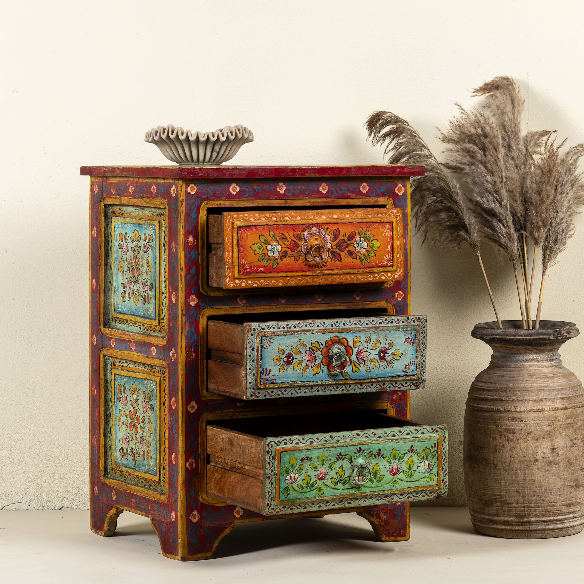 Side angle image with open drawers of a  hand-painted reclaimed wood nightstand with multiple drawers, featuring vibrant colours and floral patterns, placed next to a vase with dried plants.