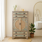 Lifestyle imagination of a hand-painted cabinet made from mango wood, featuring rattan inlay doors framed by a carved circular motif, floral blue and green accents, and five drawers for additional storage.