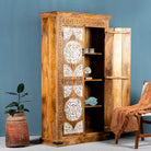 Side angle image with open door of a carved wood furniture wardrobe made from mango wood with whitewashed floral doors, wooden wardrobe in Ibiza-inspired style, 100 × 40 × 180 cm.