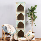 A tall hand-carved mango wood shelf with distressed pastel blue finish, featuring four open shelves, placed next to a chair with a plant and cushions.