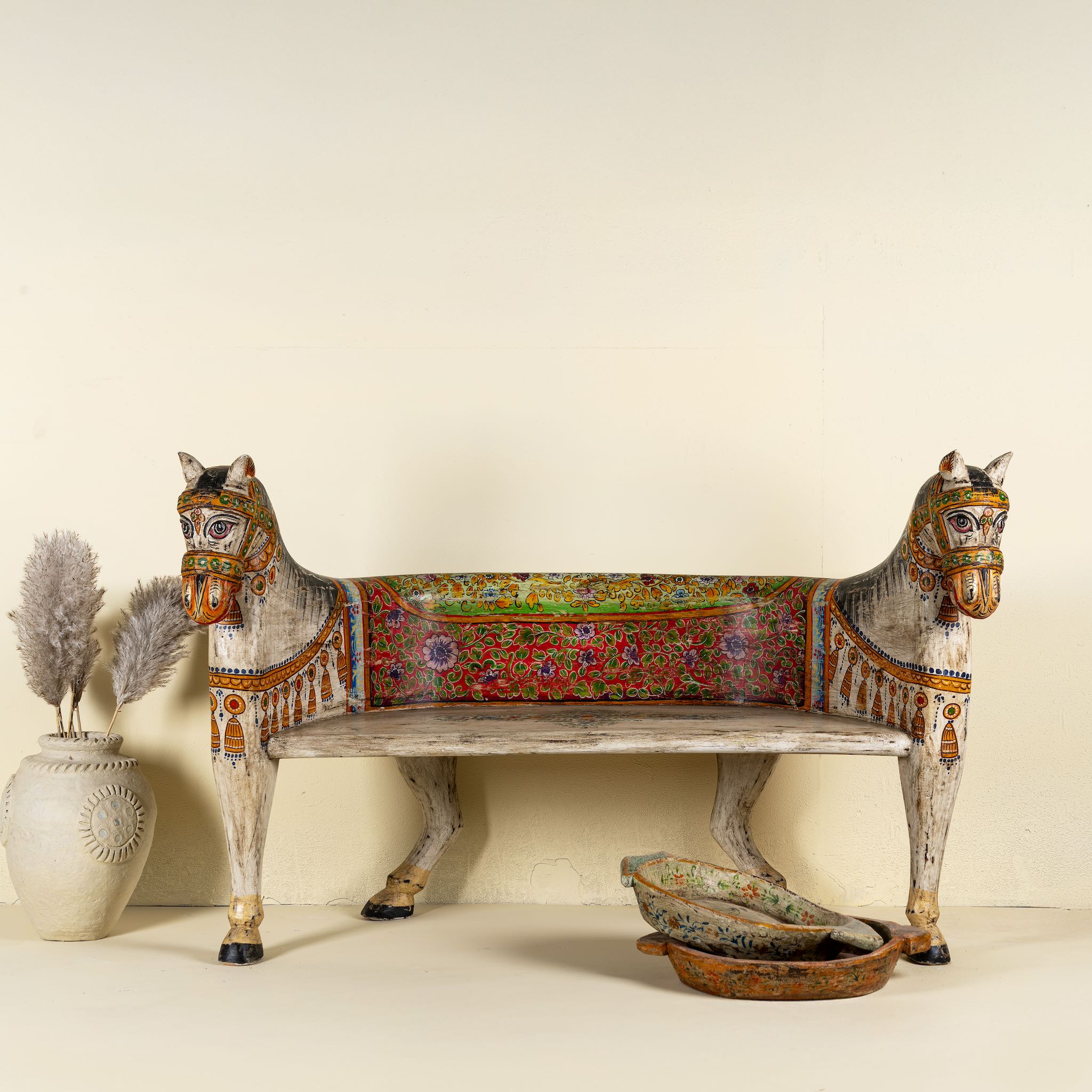 Front view image of a hand-painted bench made from reclaimed wood, designed with intricately carved double horse heads at each end, featuring a vibrant floral backrest and detailed decorative patterns, blending traditional craftsmanship with whimsical artistry.

