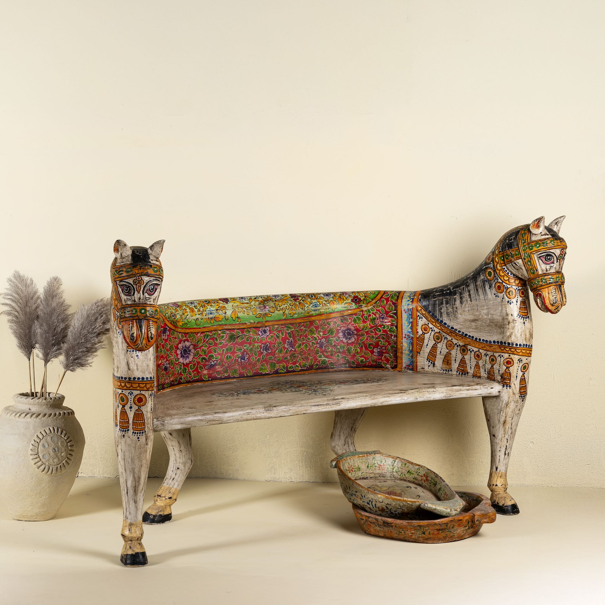 Side angle image of a hand-painted bench made from reclaimed wood, designed with intricately carved double horse heads at each end, featuring a vibrant floral backrest and detailed decorative patterns, blending traditional craftsmanship with whimsical artistry.
