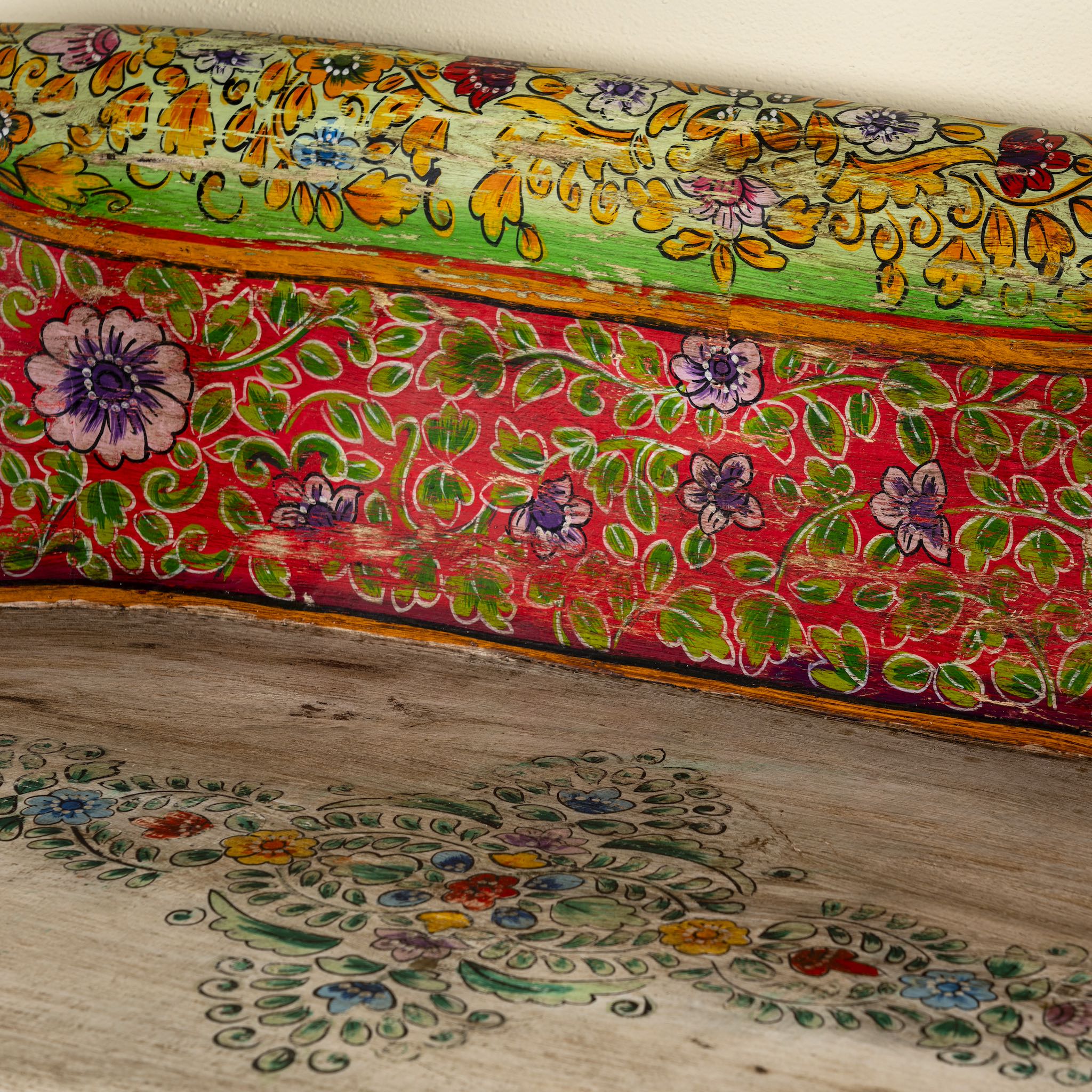 Close up image of a hand-painted bench made from reclaimed wood, designed with intricately carved double horse heads at each end, featuring a vibrant floral backrest and detailed decorative patterns, blending traditional craftsmanship with whimsical artistry.
