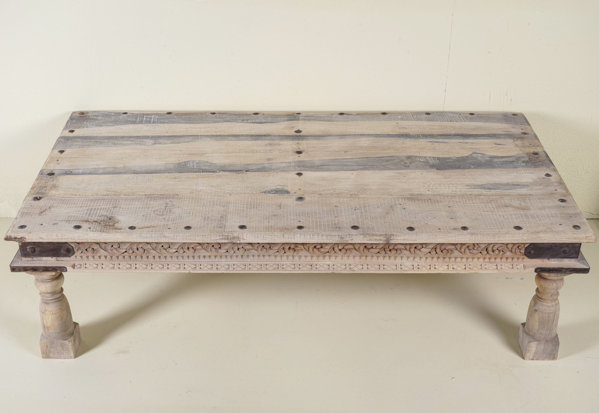 Close up of the table top of a rustic coffee table made from old reclaimed teak wood, featuring a weathered finish, turned legs, decorative carved apron, and visible metal rivets, styled with earthy decor in a minimal interior setting.
