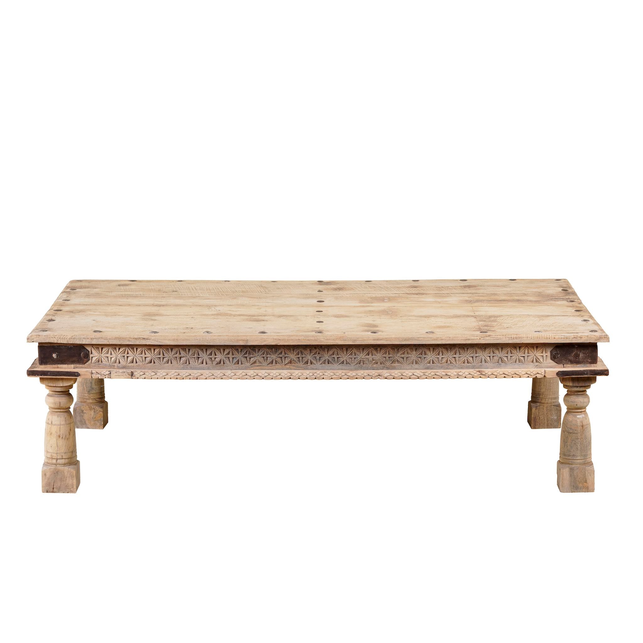 White background image of a rustic coffee table made from old reclaimed teak wood, featuring a weathered finish, turned legs, decorative carved apron, and visible metal rivets, styled with earthy decor in a minimal interior setting.
