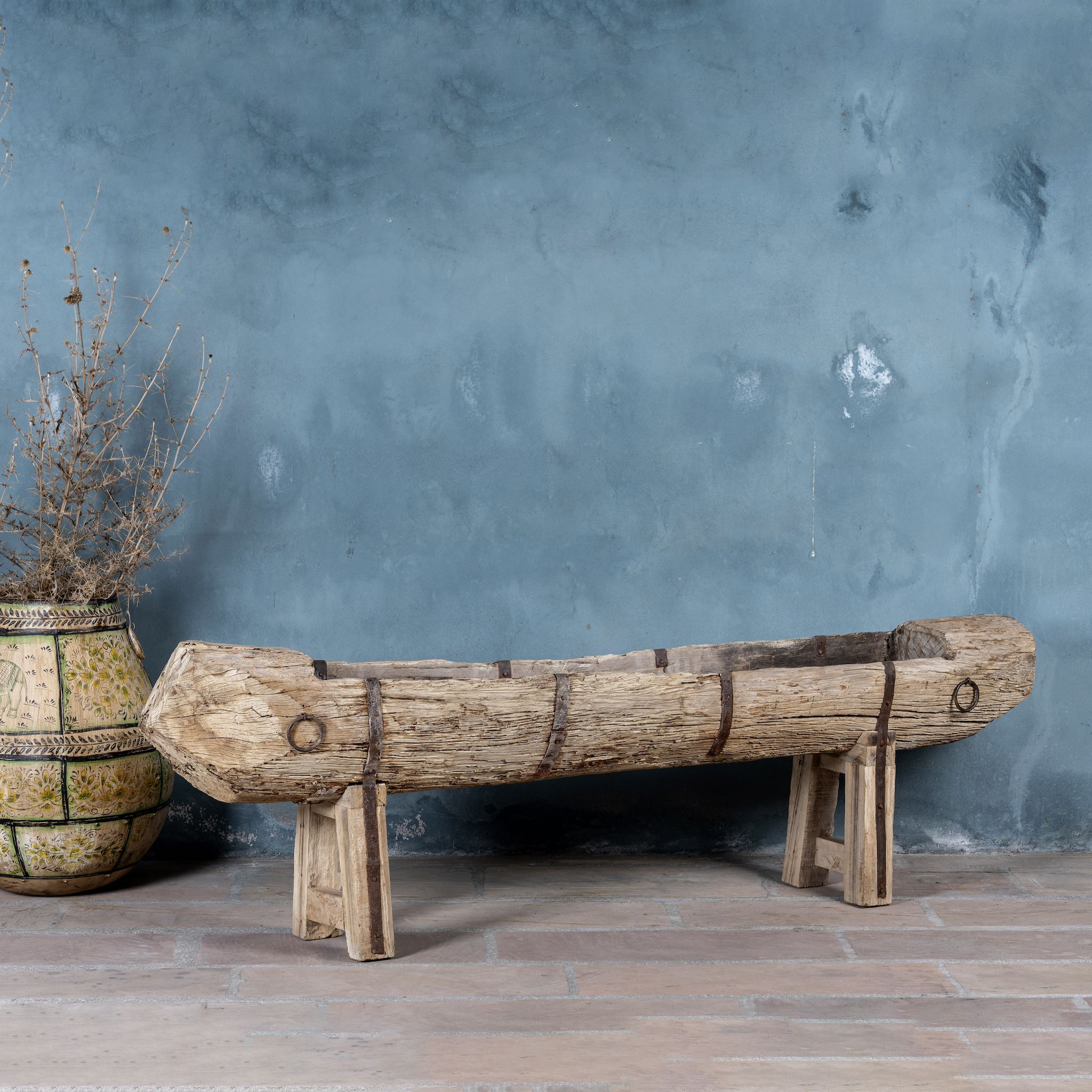 Wooden log bench against a textured blue wall with a plant on the left.