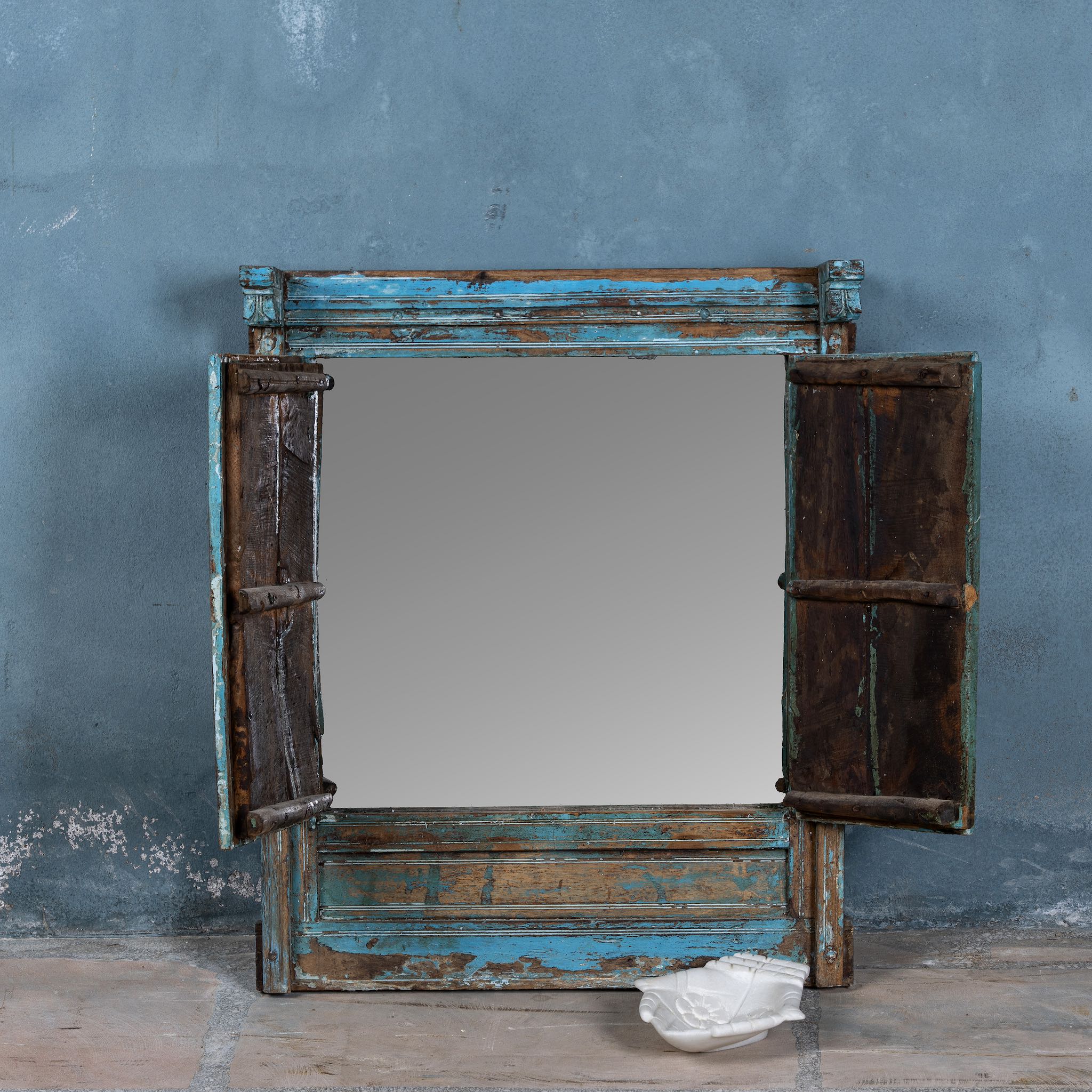 Vintage-style mirror with wooden frame and peeling blue paint against a textured gray wall.