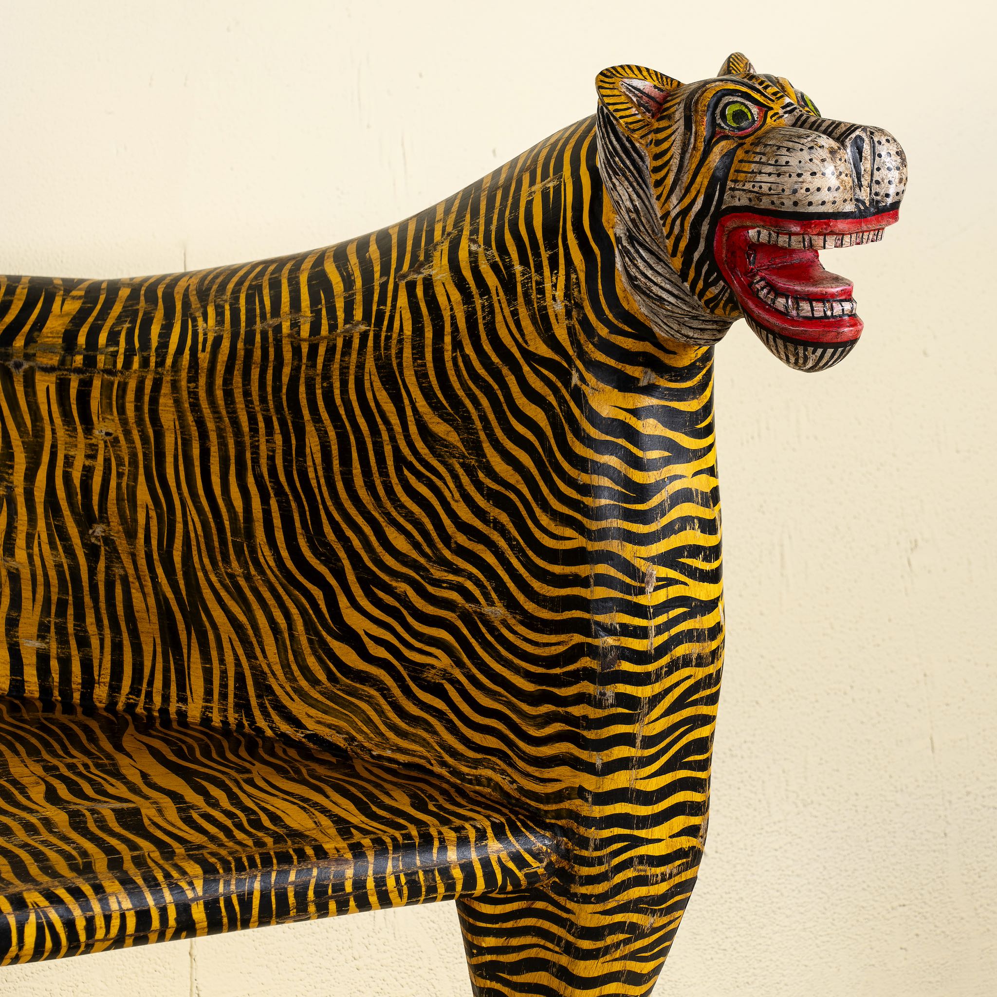 Close up image of the head of a bold hand-painted bench made from reclaimed wood, designed with double tiger heads at each end and a vibrant yellow-and-black striped pattern throughout, combining whimsical animal form with striking folk-art flair.
