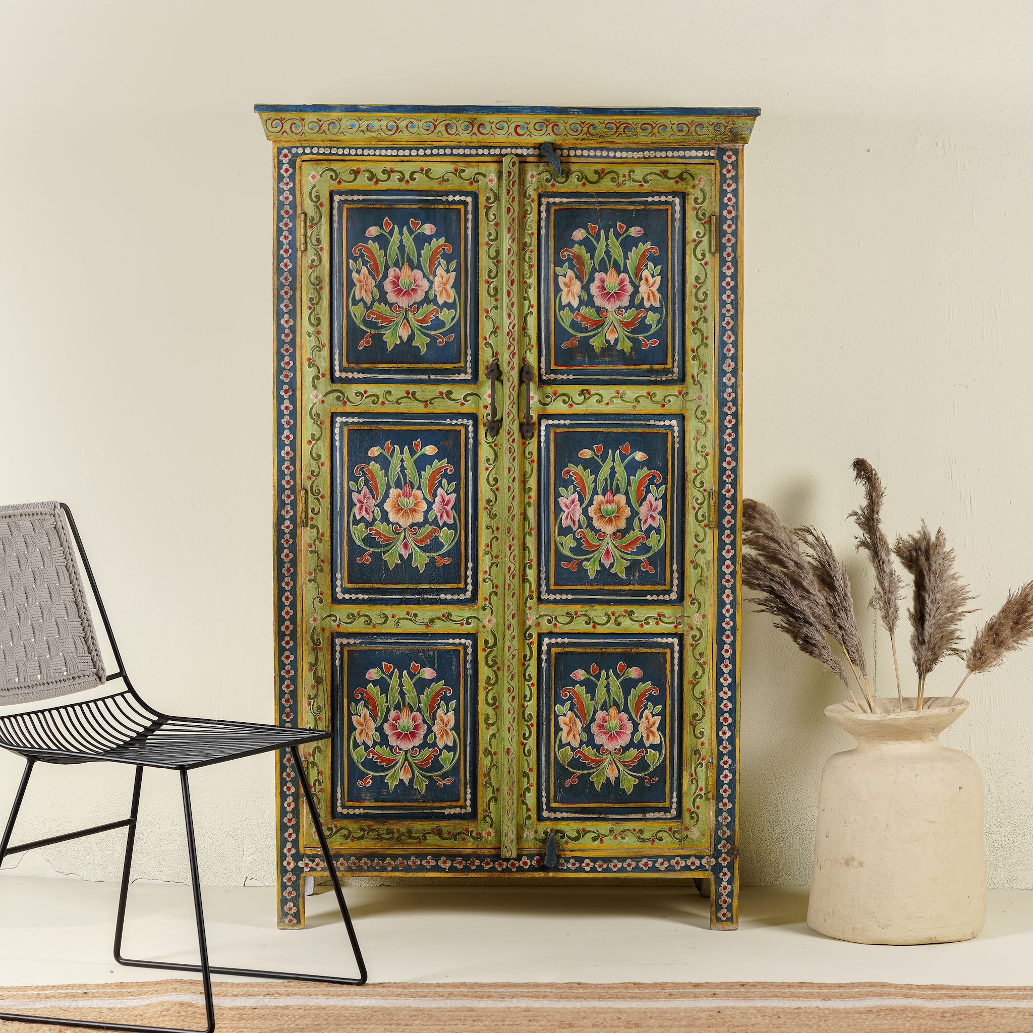 A vibrantly hand-painted reclaimed wood vintage cabinet with floral motifs, in a colorful and traditional design, standing in a room with a chair and a decorative vase beside it.