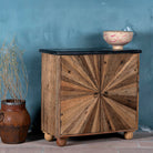 Side angle image of a rustic-style cabinet made from reclaimed wood, featuring a striking sunburst pattern on the double doors, round metal pulls, and a contrasting black marble top that adds a refined touch to its natural, weathered look.
