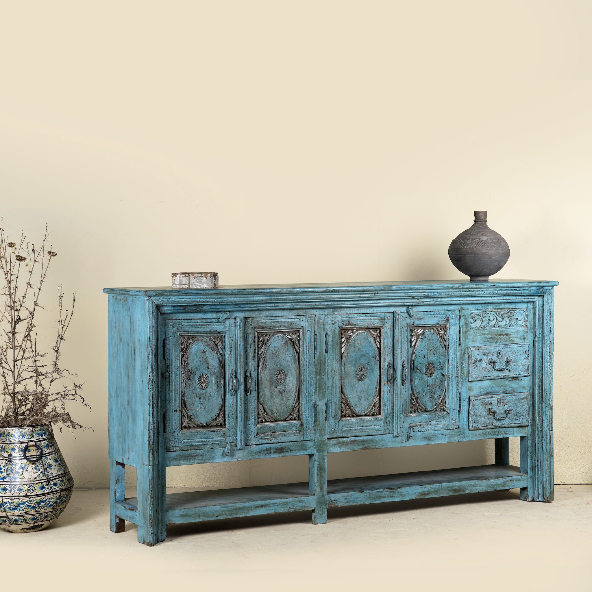 Side angle picture of an antique console table made from solid teak wood, featuring four carved doors, three drawers, and a striking distressed turquoise finish for bold vintage flair.