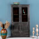 A shabby chic-style reclaimed teak wood showcase cabinet with glass doors and various decorative items displayed on its shelves, set against a plain blue wall.