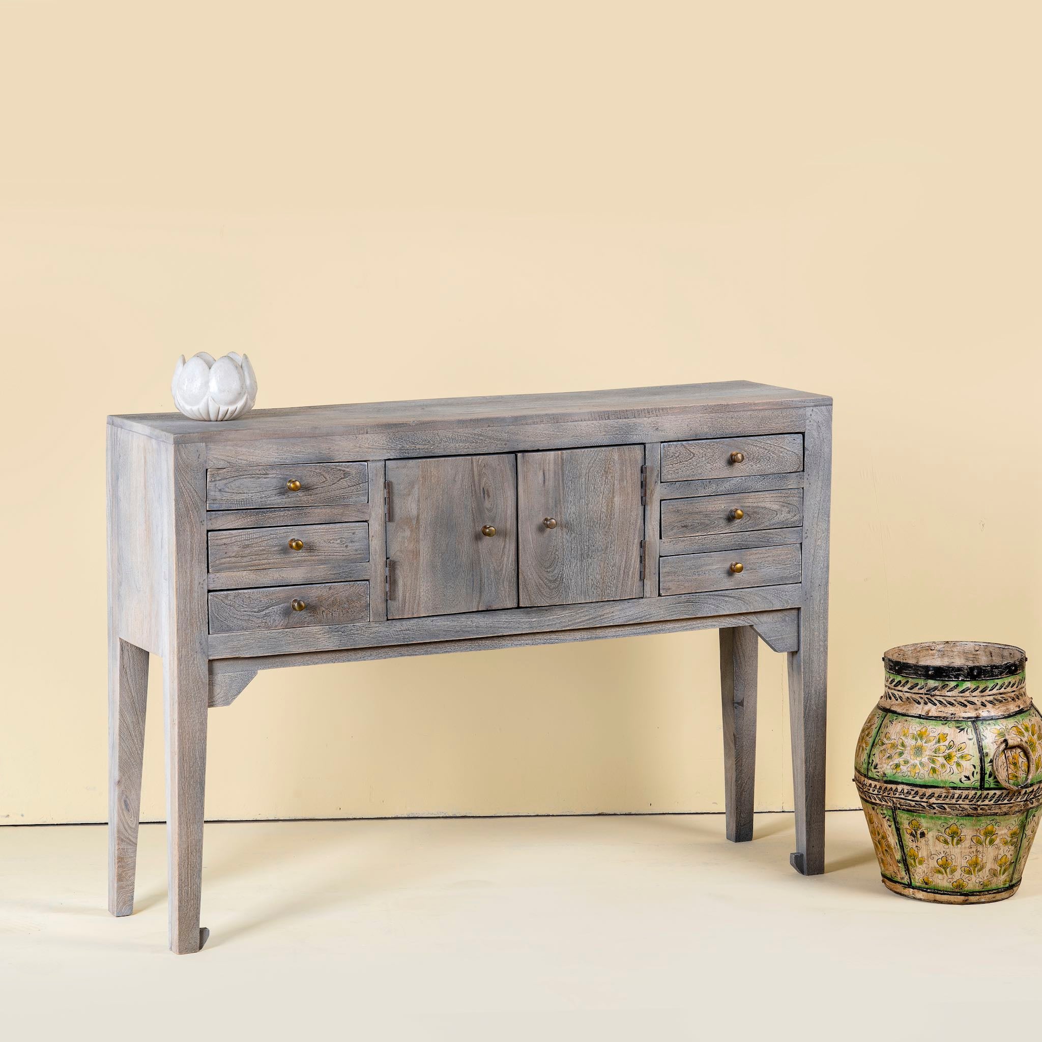 Elegant console table made from solid mango wood, featuring a weathered grey finish, six drawers, and two central doors with brass knobs.