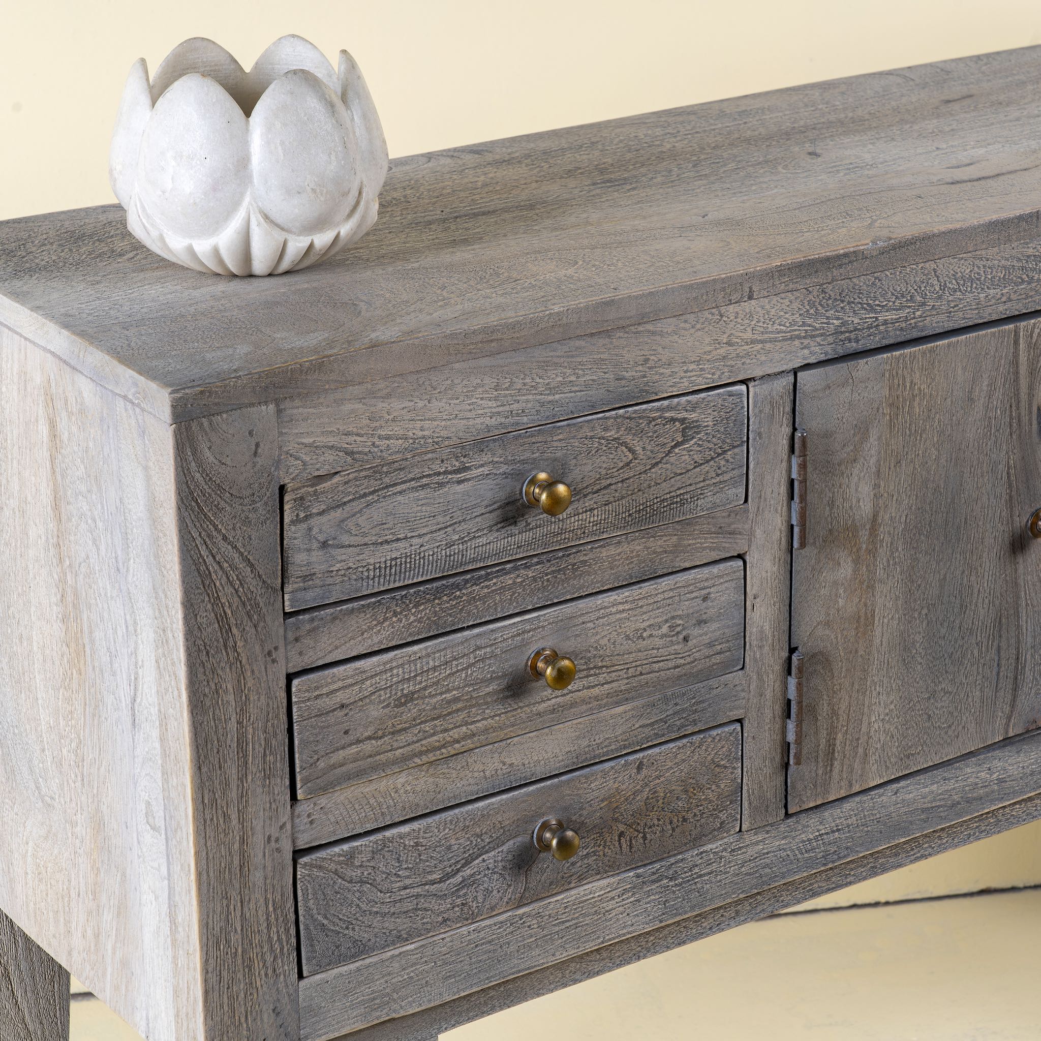 Close up picture of a elegant console table made from solid mango wood, featuring a weathered grey finish, six drawers, and two central doors with brass knobs.