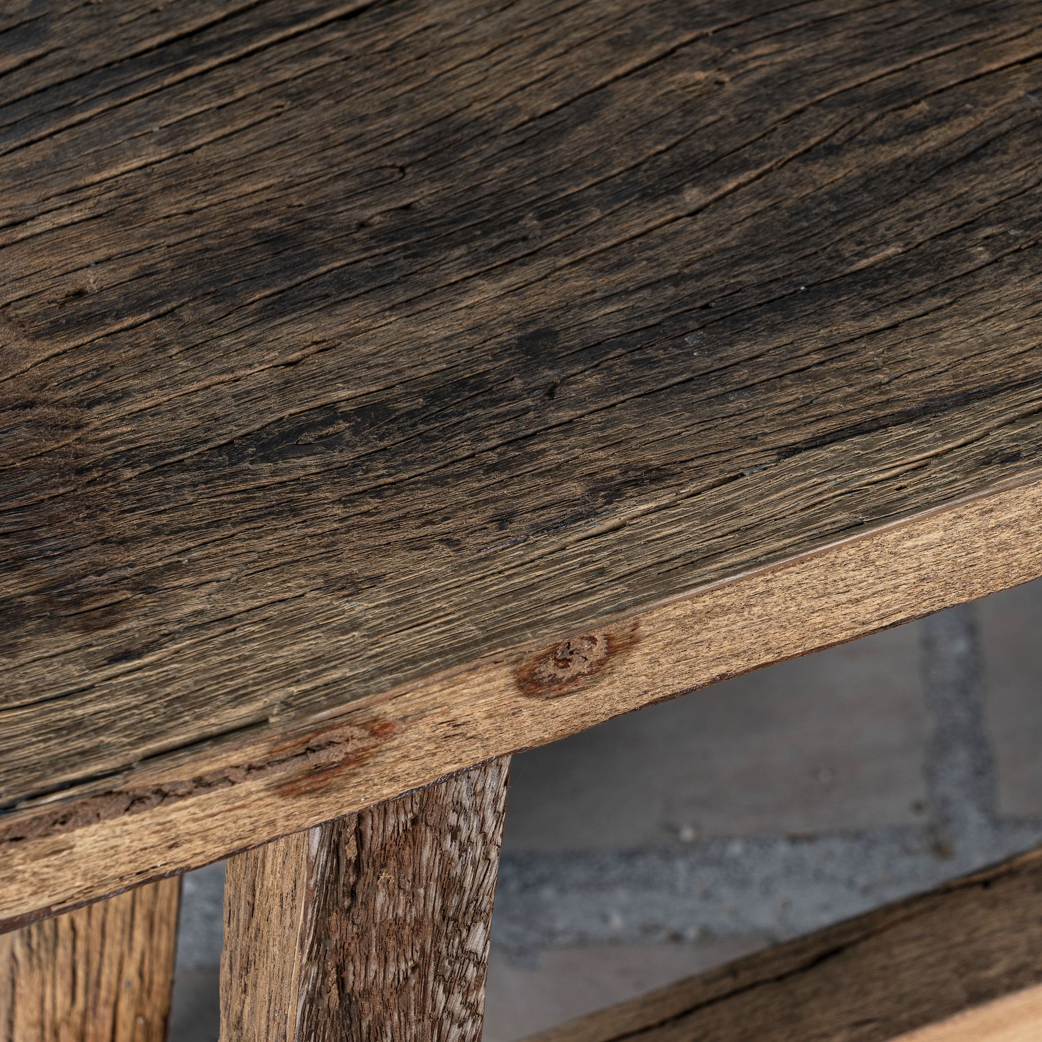 Close up picture of a rustic wooden bench made from reclaimed wood, featuring a long, narrow seat with a weathered finish and sturdy angled legs connected by a simple support beam.
