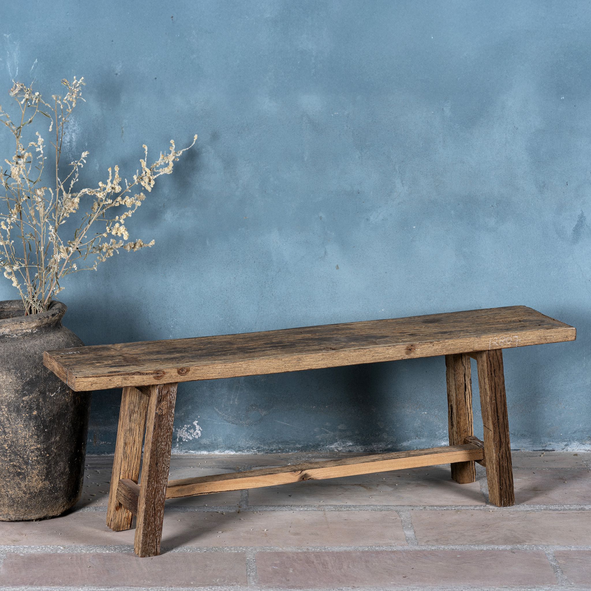 Front view image of a rustic wooden bench made from reclaimed wood, featuring a long, narrow seat with a weathered finish and sturdy angled legs connected by a simple support beam.
