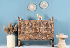 An antique Indian treasure chest with sandblasted surface and iron applications, featuring intricate hand-carved patterns, displayed against a blue wall with decorative items around it.