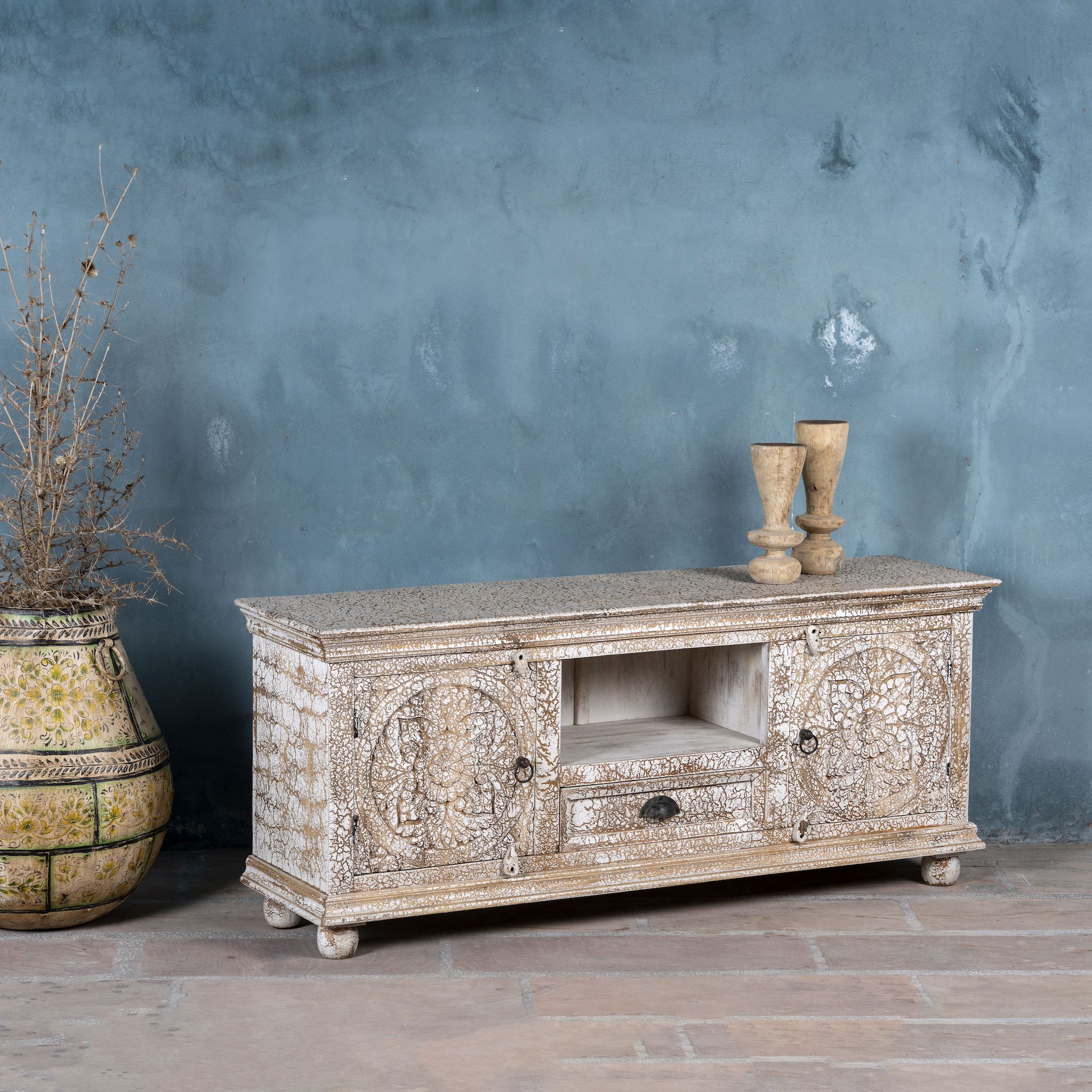 Side angle image of a carved mango wood lowboard with floral doors, crackled vintage finish, drawer, and open shelf, 150 × 40 × 64 cm.