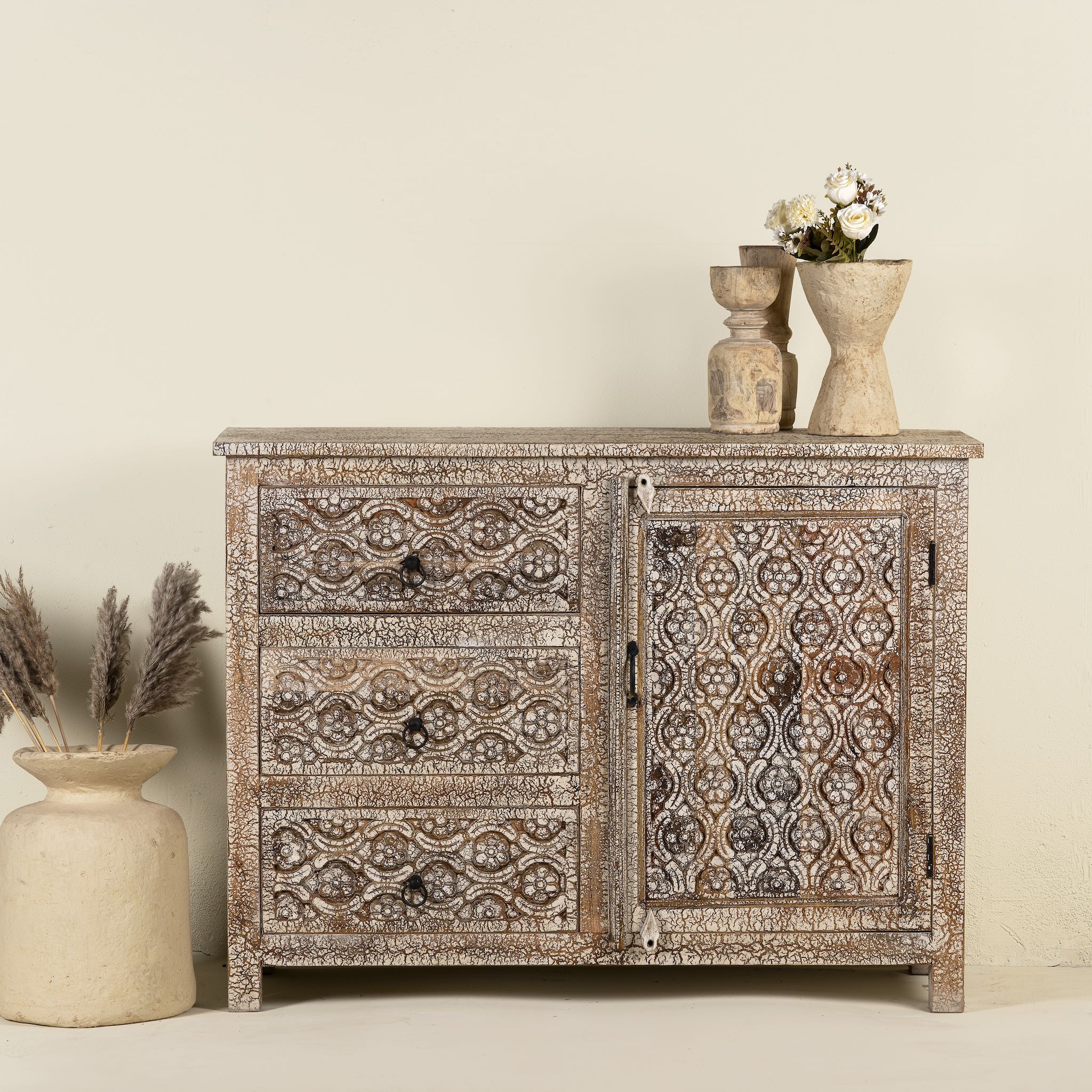 Front view image of a hand-carved sideboard made from mango wood, featuring intricate geometric and floral patterns across the drawers and cabinet door, with a distressed whitewashed finish that enhances its vintage artisanal appeal.
