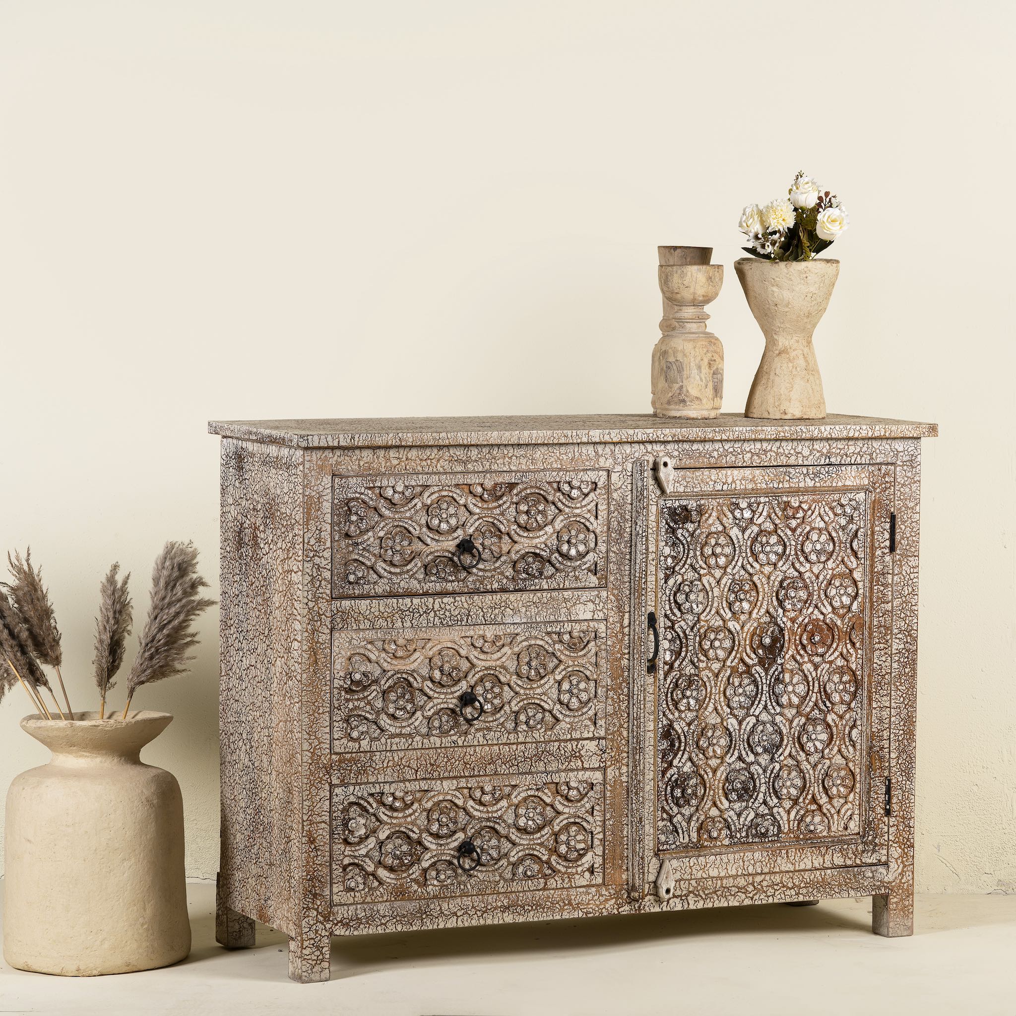 Side angle image of a hand-carved sideboard made from mango wood, featuring intricate geometric and floral patterns across the drawers and cabinet door, with a distressed whitewashed finish that enhances its vintage artisanal appeal.
