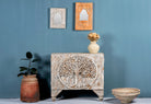 A beige mango wood cupboard with intricate hand-carved tree designs on the front, with a framed mirror above, a potted plant to the left, and a decorative bowl to the right on a blue background.