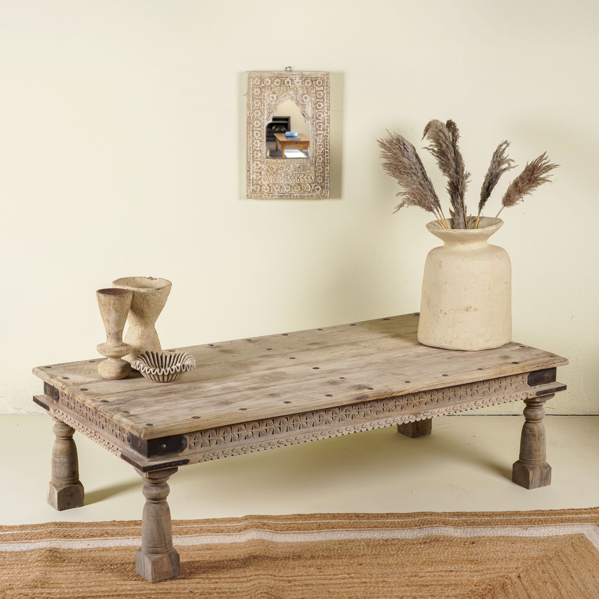 Side angle of rustic coffee table Ritvik made from old reclaimed teak wood, featuring a weathered finish, turned legs, decorative carved apron, and visible metal rivets, styled with earthy decor in a minimal interior setting.

