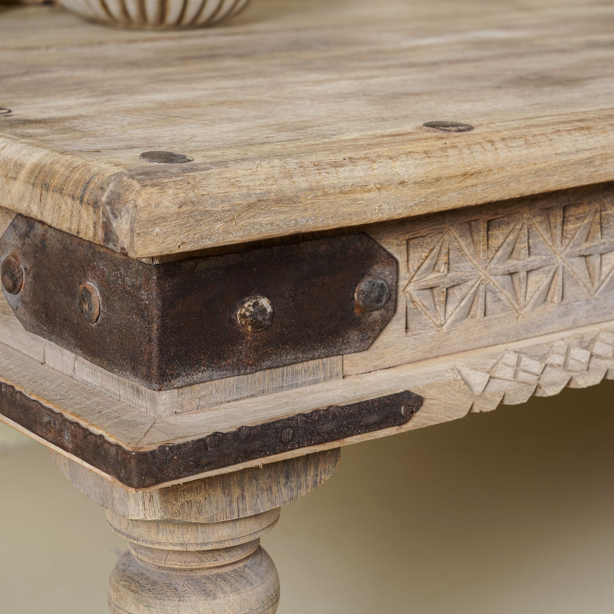 Close up of a corner of rustic coffee table Ritvik made from old reclaimed teak wood, featuring a weathered finish, turned legs, decorative carved apron, and visible metal rivets, styled with earthy decor in a minimal interior setting.
 