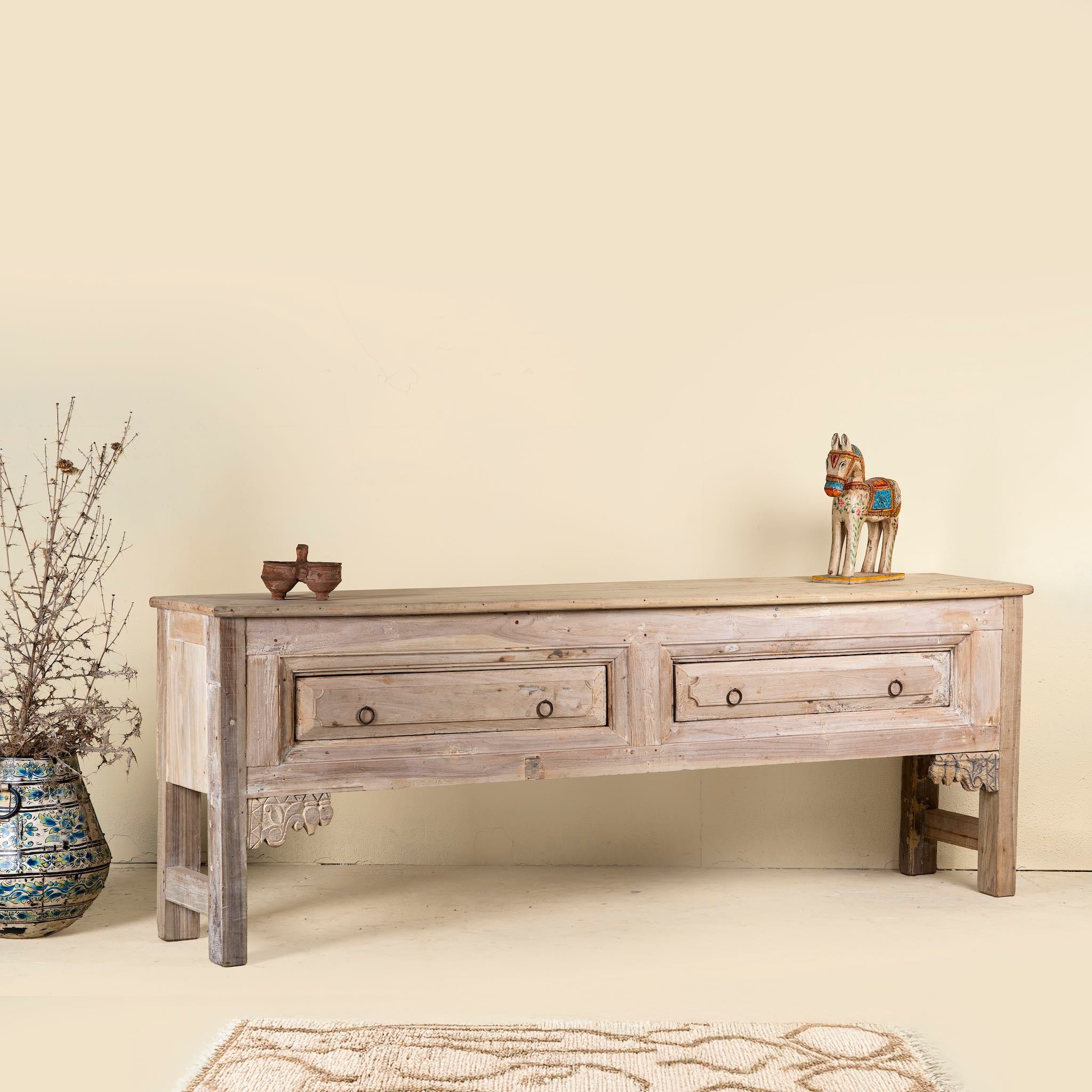 Side angle picture of a vintage-style teak console table with two spacious drawers, simple carved brackets, and a natural bleached wood finish for a clean rustic look.