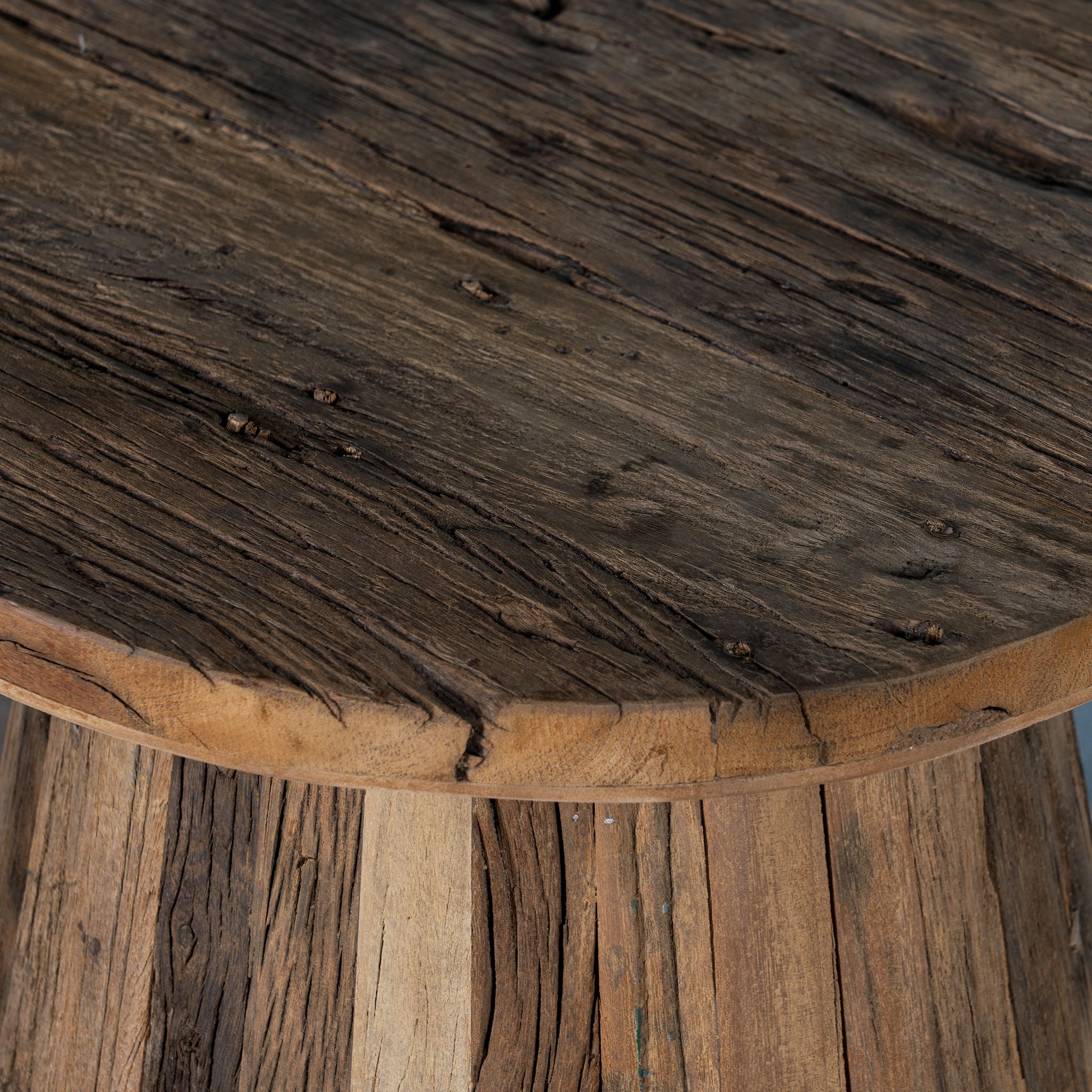 Close up image of a rustic-style coffee table crafted from recycled wood, featuring a round top and tapered conical base with a natural, weathered finish showcasing varied wood tones and textures.