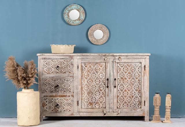 A hand-carved mango wood sideboard with a distressed finish, featuring floral lattice carvings, multiple storage compartments, and placed against a blue wall with decorative items around it.