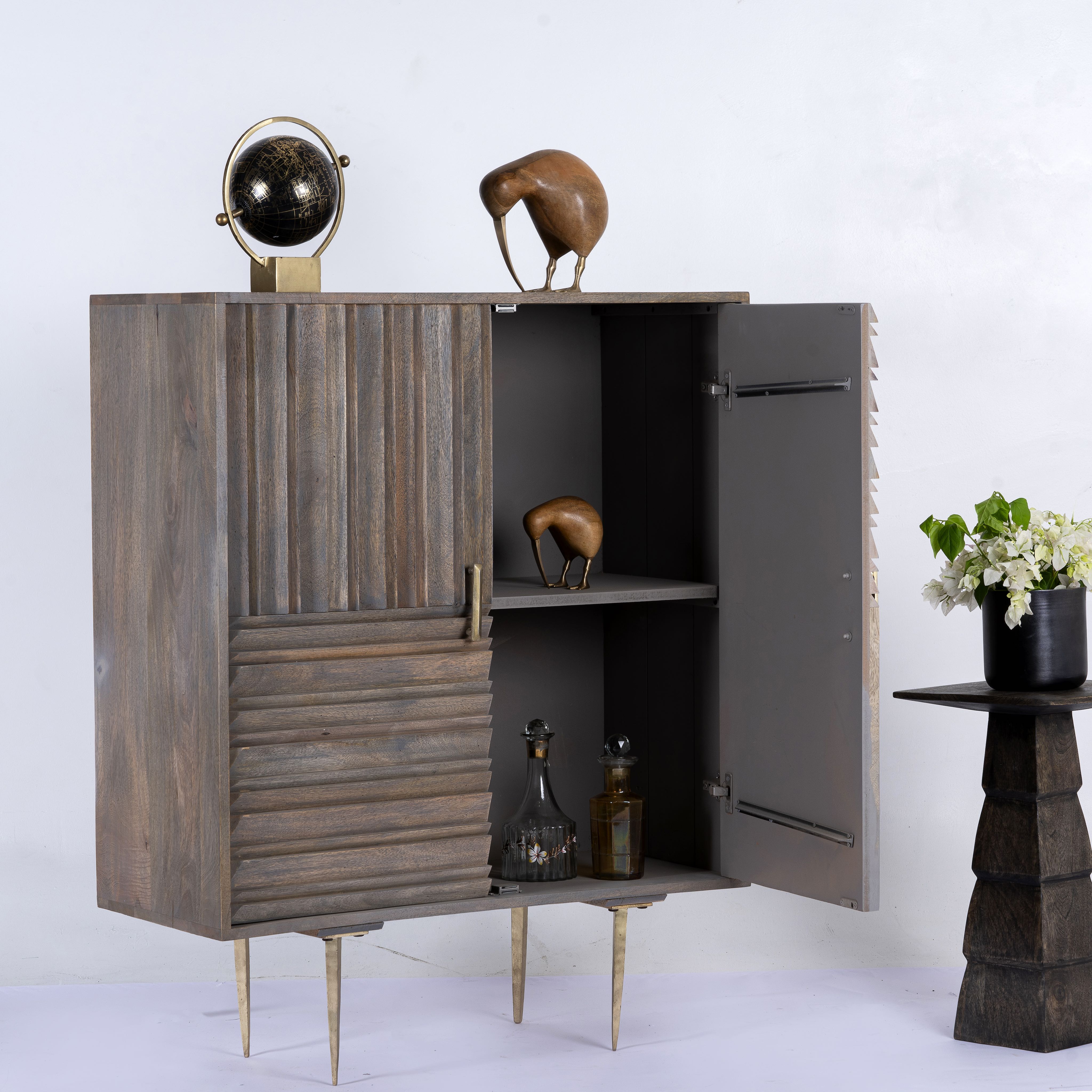 Side angle image with open door of a Grey carved cabinet in mango wood with linear carvings and brass‑plated iron base, 92 × 42 × 115 cm.