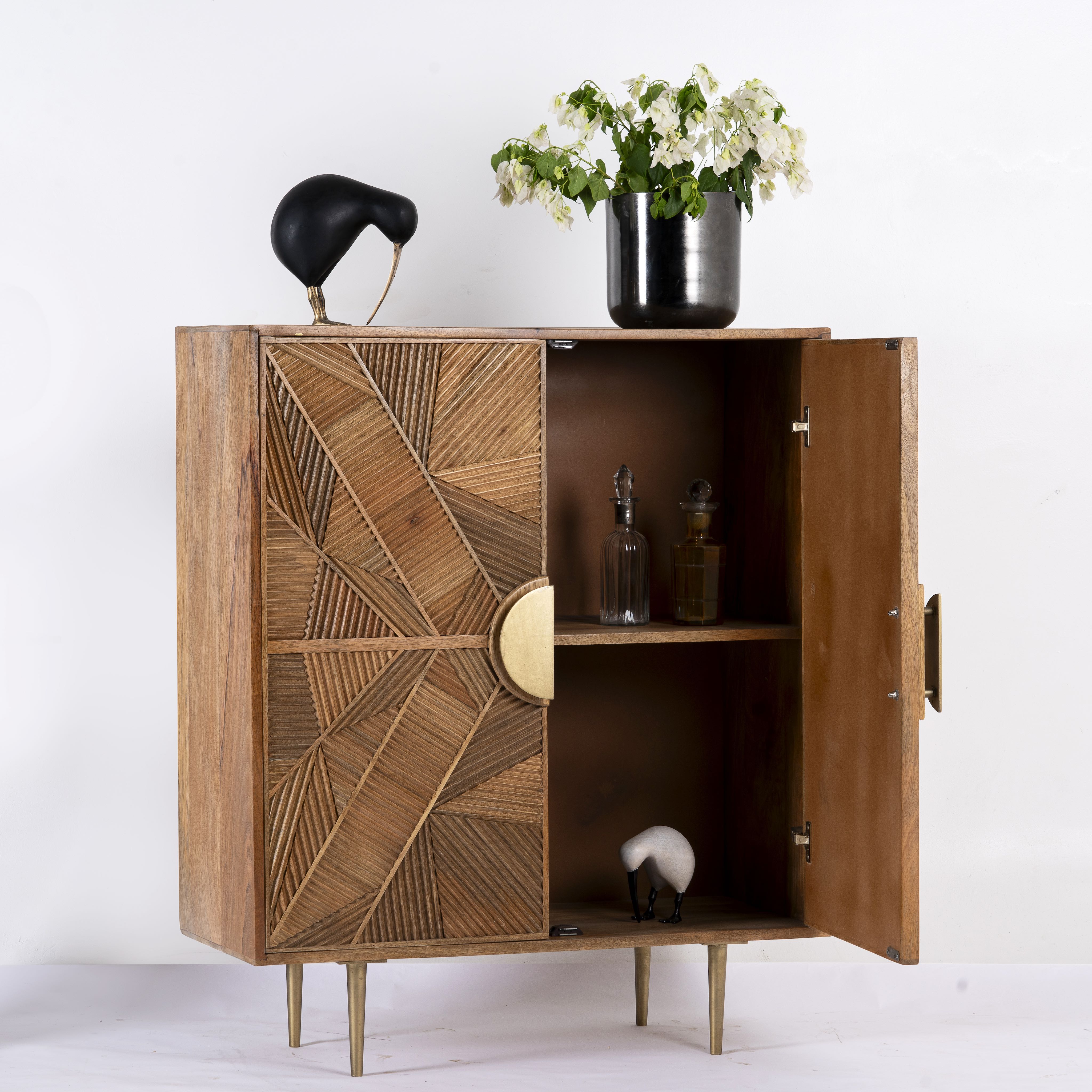 Side angle image with open doors of a natural mango wood cupboard with carved linear design and antique brass legs, 91 × 40 × 109 cm.