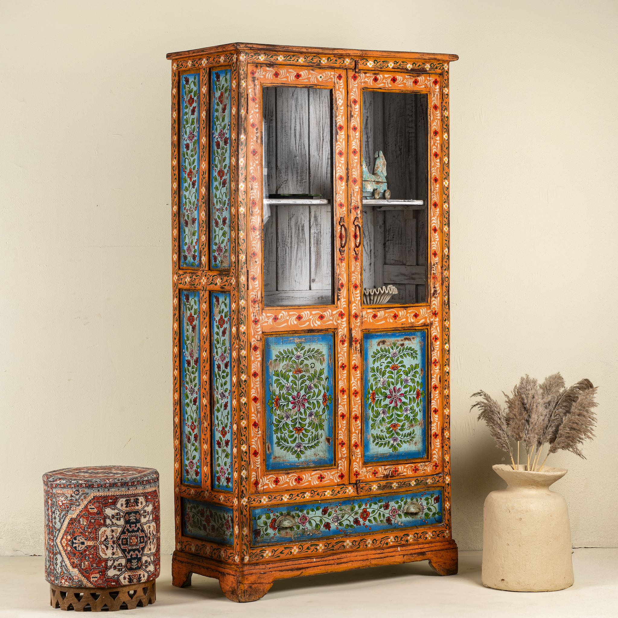 Side angle image of a hand-painted teak wood showcase with floral motifs and glass doors – sustainable furniture crafted from recycled wood with traditional Indian artistry.