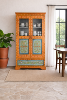 Lifestyle imagination of a of a hand-painted teak wood showcase with floral motifs and glass doors – sustainable furniture crafted from recycled wood with traditional Indian artistry.