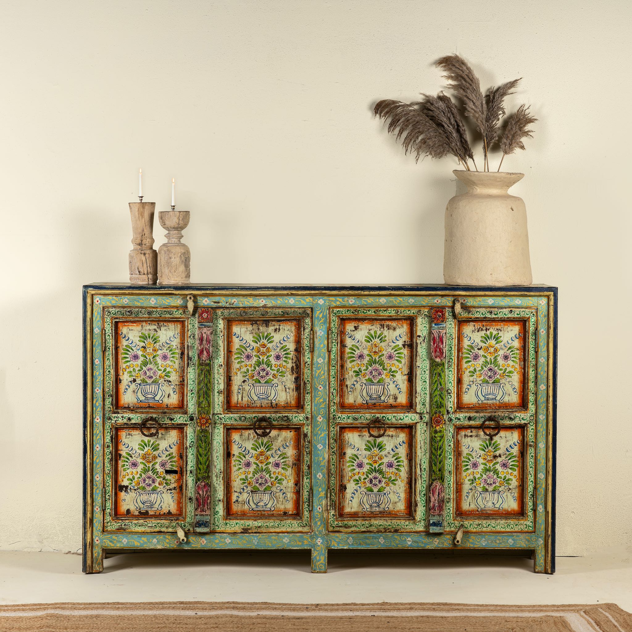 Hand-painted sideboard made from reclaimed wood, showcasing vibrant floral motifs in green, pink, yellow, and blue on a distressed turquoise base, with six panelled doors and rustic iron ring handles.