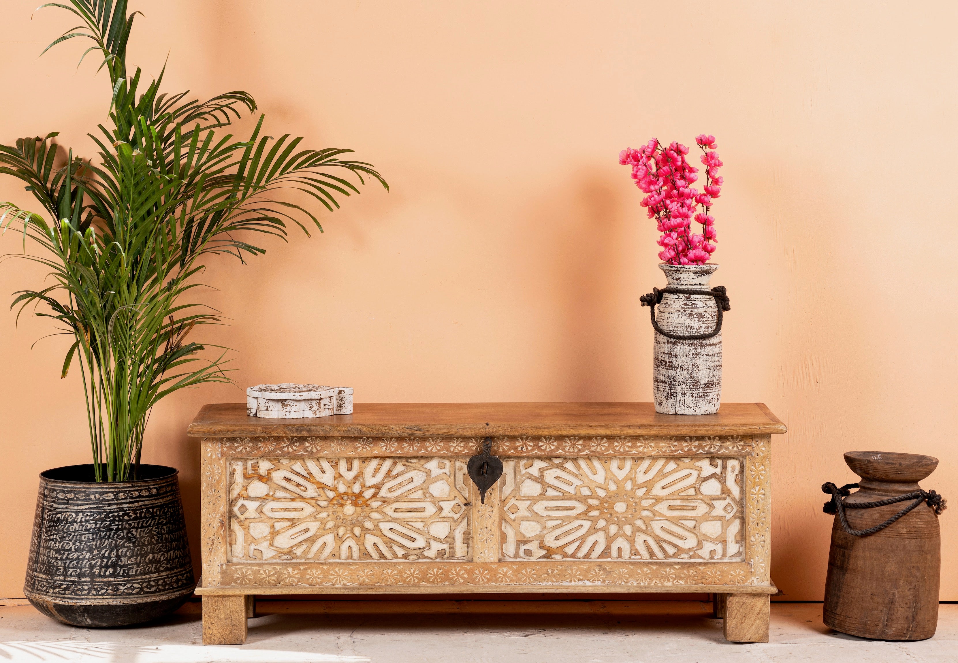 A wooden storage chest with intricate carvings on the front, placed on a floor against a wall, with a plant to the left and a decorative vase on top of the chest.