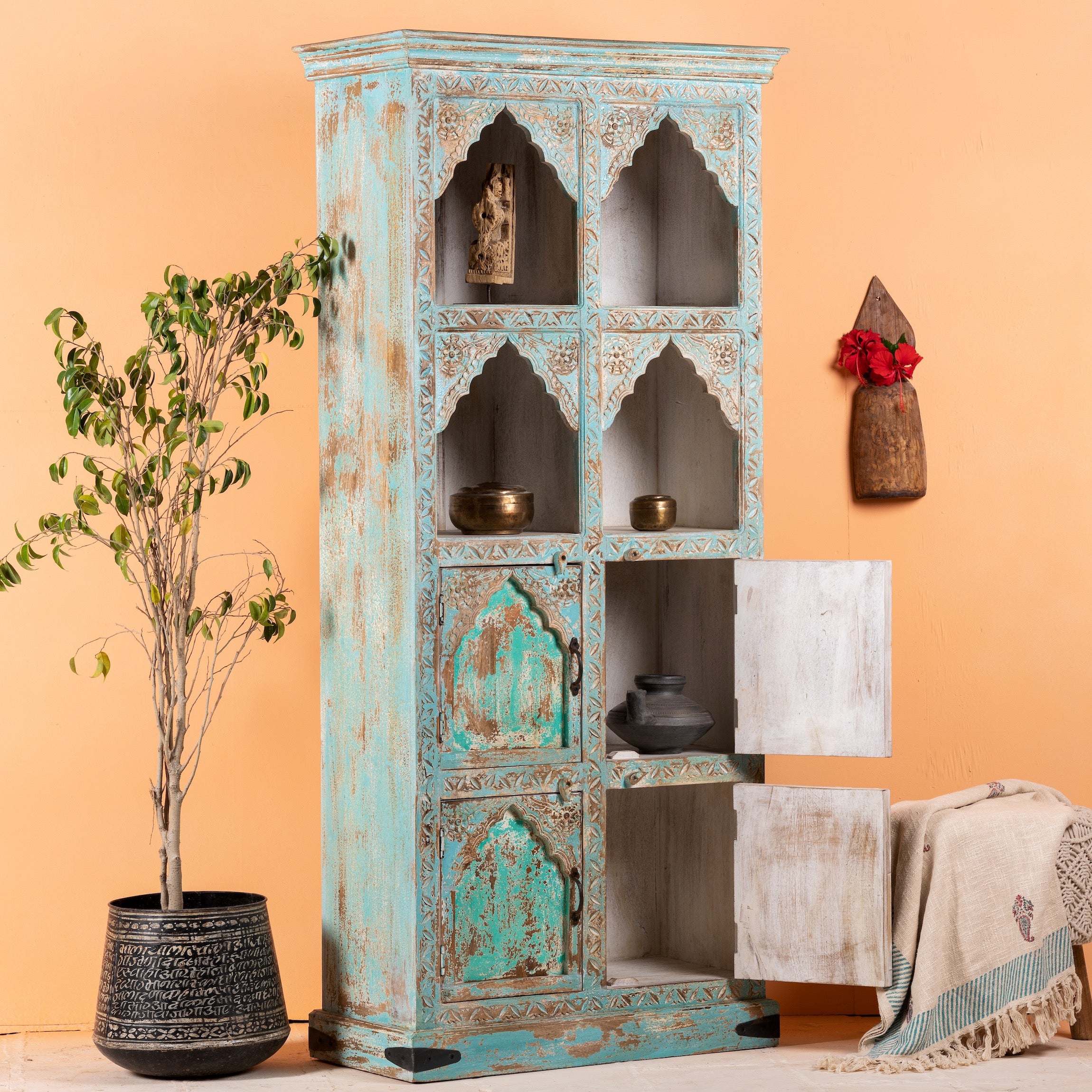 Side angle image with open doors of a hand-carved mango wood shelf with arched open display and lower cabinets – vintage home decoration piece in distressed pastel blue finish with Indian architectural detailing.