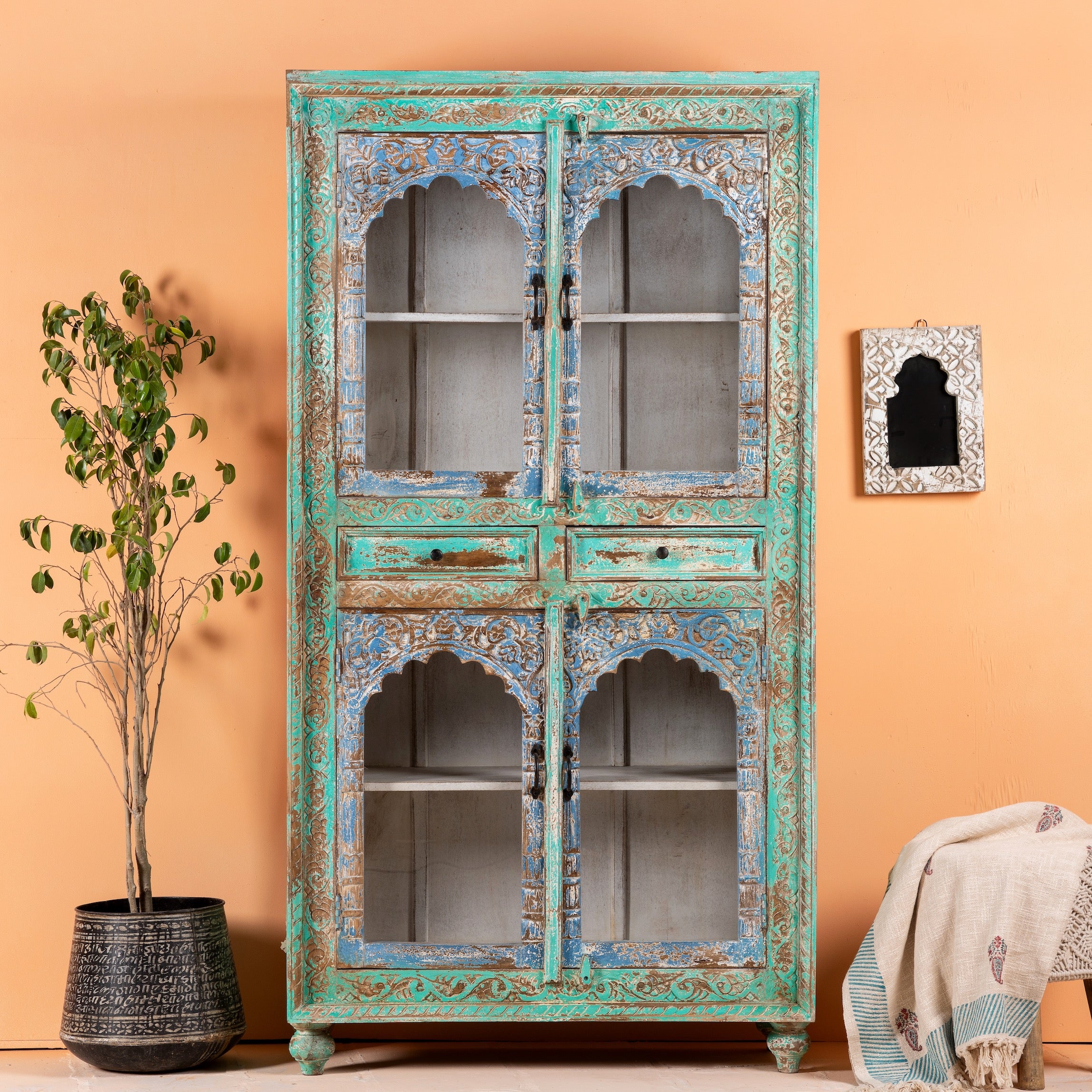 A vintage hand-carved mango wood showcase with distressed turquoise and blue finish, featuring glass panelled doors and wooden carvings, placed in a room with a plant and decorative items.
