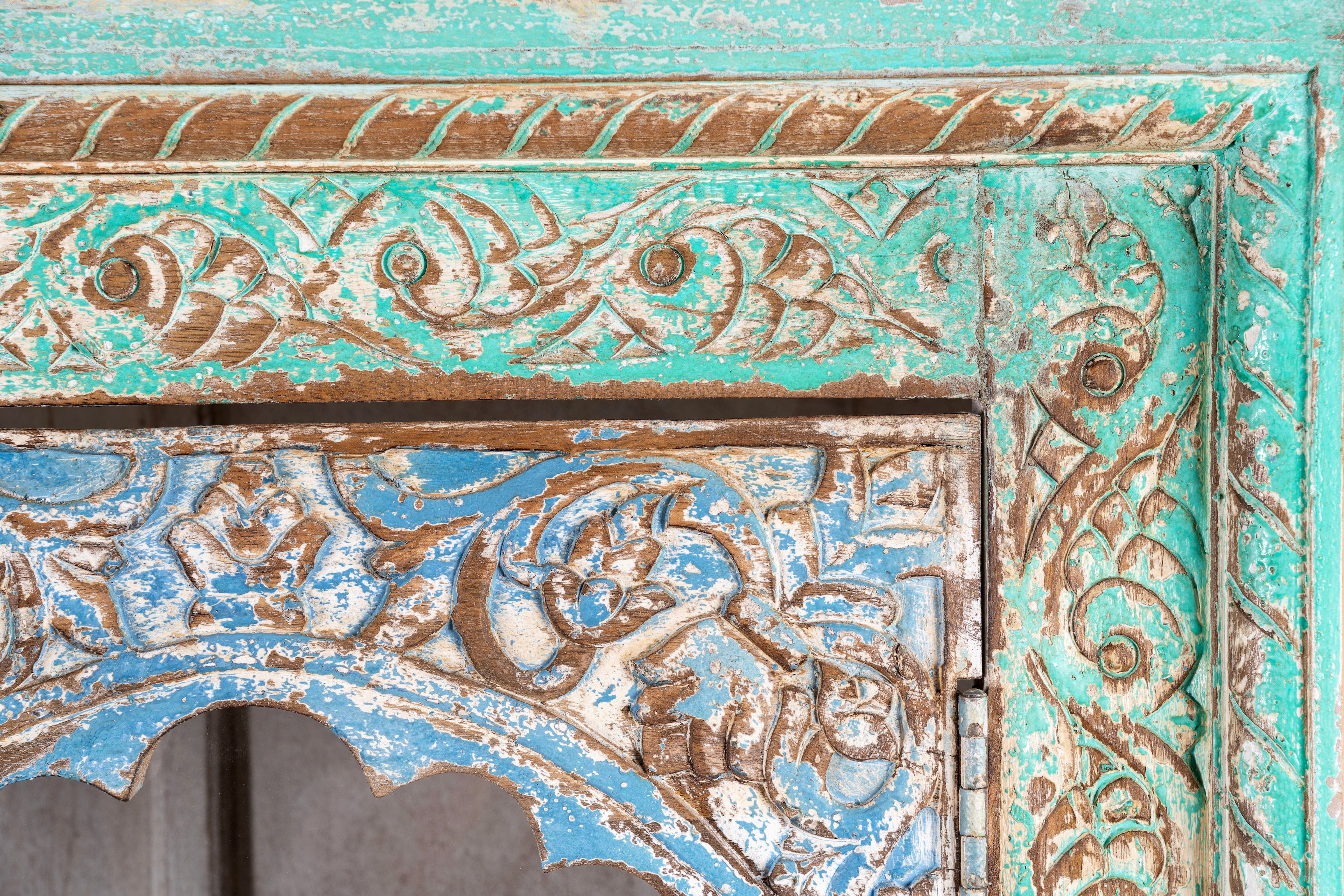 Close up image of a hand-carved mango wood showcase with glass doors and drawers – carved wooden furniture in distressed turquoise and blue, combining artisan detail with vintage charm.